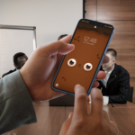 Par de manos sosteniendo el móvil con el fondo de pantalla de Wachi en uso, con una vista de una oficina y personas trabajando al fondo.