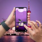 Dispositivo con un par de manos usando el nuevo fondo de pantalla de Wachi, donde se muestra a Wachi brujita volando sobre su escoba mágica en un bosque encantado.