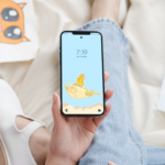 Imagen de una chica sonriente utilizando su teléfono móvil con el fondo de pantalla del pollito chibi volando en un avión.